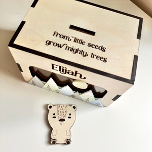 May include: Wooden box with a slot on top for coins. The box has a cutout on the front with a mountain scene and the words "From little seeds grow mighty trees". The box is personalized with the name "Elijah". A small wooden bear figurine is in front of the box.