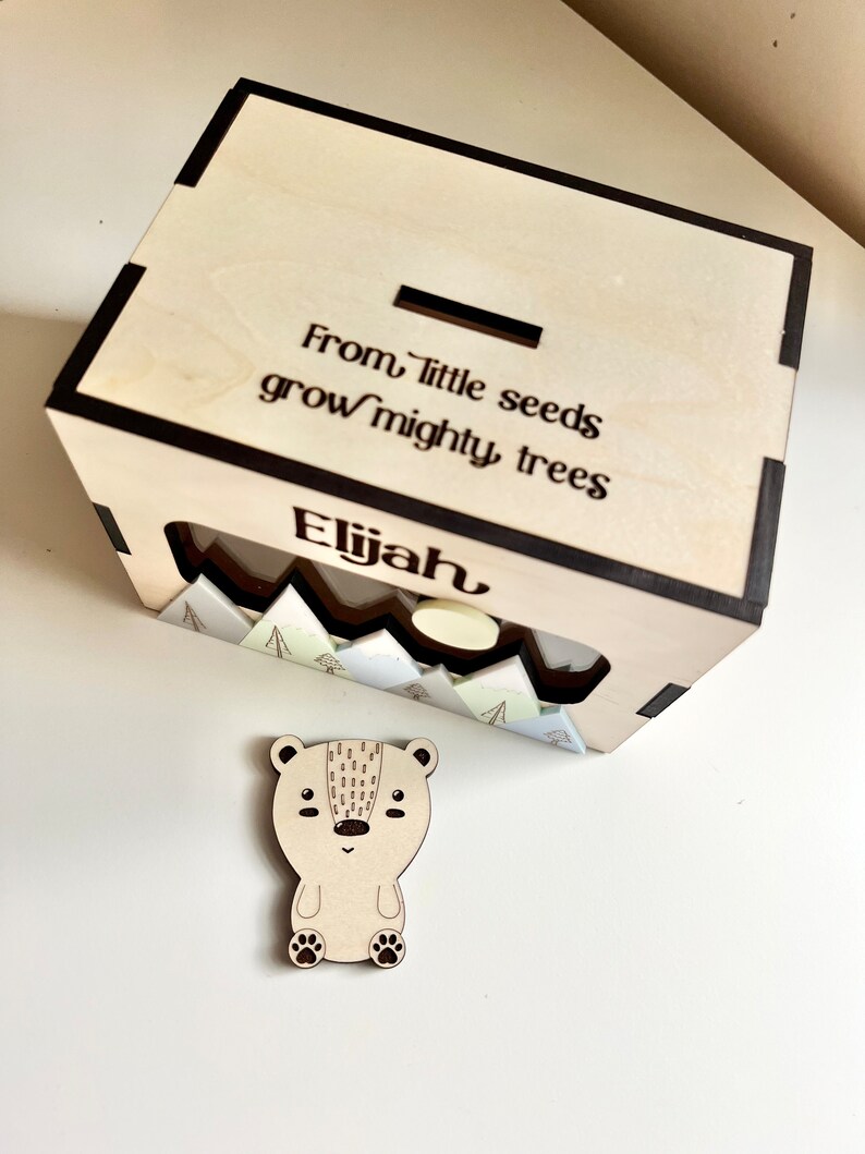 May include: Wooden box with a slot on top for coins. The box has a cutout on the front with a mountain scene and the words "From little seeds grow mighty trees". The box is personalized with the name "Elijah". A small wooden bear figurine is in front of the box.