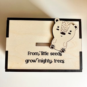 May include: Wooden box with a black frame and a small wooden bear figurine on the top. The box has a slot on the front and the text "From little seeds grow mighty trees" is engraved on the front.