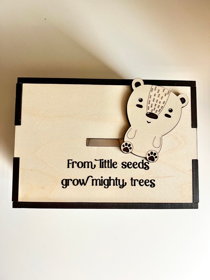 May include: Wooden box with a black frame and a small wooden bear figurine on the top. The box has a slot on the front and the text "From little seeds grow mighty trees" is engraved on the front.