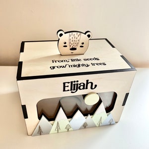May include: Wooden box with a cutout window and a small wooden bear on top. The box has a mountain scene at the bottom and the text "From little seeds grow mighty trees Elijah" on the front.