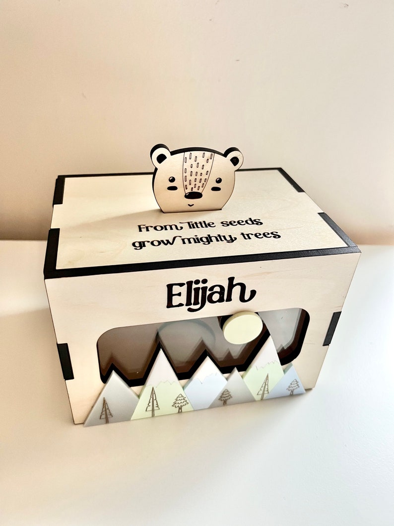 May include: Wooden box with a cutout window and a small wooden bear on top. The box has a mountain scene at the bottom and the text "From little seeds grow mighty trees Elijah" on the front.