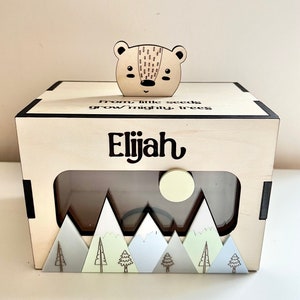 May include: A wooden box with a cutout window, featuring a mountain scene with trees and a moon. The box has a bear head on top and the text "From little seeds grow mighty trees Elijah" is engraved on the front.