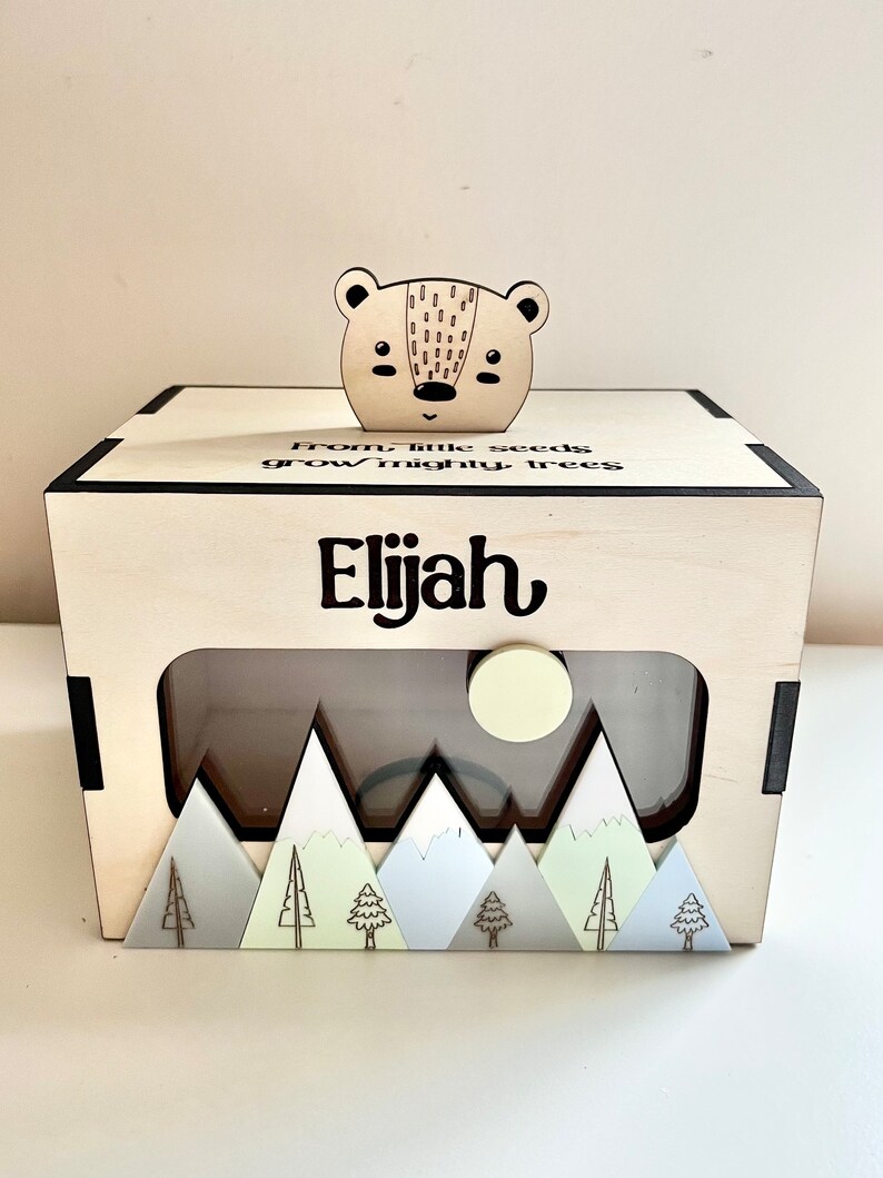May include: A wooden box with a cutout window, featuring a mountain scene with trees and a moon. The box has a bear head on top and the text "From little seeds grow mighty trees Elijah" is engraved on the front.