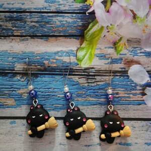 Könnte beinhalten: Drei schwarze Katzenanhänger mit rosa Wangen und einem schwarzen Besen. Jeder Anhänger ist an einer silbernen Kette mit einer blauen, weißen oder silbernen Perle befestigt.