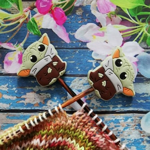 Könnte beinhalten: Zwei grüne, braune und weiße Maschenmarkierer in Form von Charakteren mit Stricknadeln aus Holz. Die Nadeln sind in ein farbenfrohes, teilweise gestricktes Projekt eingesetzt. Der Hintergrund zeigt ein Blumenmuster auf einer blauen Holzoberfläche.