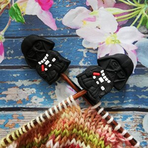 Könnte beinhalten: Zwei schwarze Darth Vader Strickmarkierer auf Holzstricknadeln mit einem mehrfarbigen gestrickten Stoff im Hintergrund.