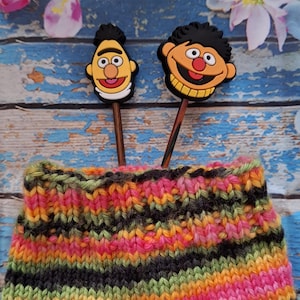 Könnte beinhalten: Ein farbenfrohes Strickstück mit einer lebendigen Mischung aus Regenbogenfarben, darunter Rosa, Orange, Grün und Lila. Zwei Topper mit Charaktermotiven sind in das Strickstück eingesetzt. Der Hintergrund zeigt eine verwitterte blaue Holzoberfläche und florale Akzente.