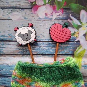 Könnte beinhalten: Nahaufnahme eines grünen und mehrfarbigen Strickartikels mit zwei dekorativen Stricknadelaufsätzen. Einer ist ein weißes Schaf mit rosa Nadeln, der andere ein rosa Wollknäuel mit einer grünen Nadel. Der Hintergrund ist ein Blumendesign.