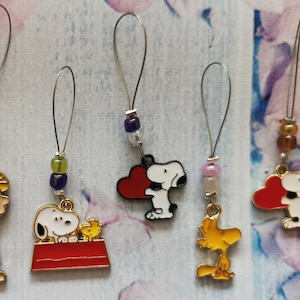 Könnte beinhalten: Sechs bunte Perlen-Maschenmarkierer mit verschiedenen Peanuts-Charakteren, darunter Snoopy, Woodstock und Lucy. Die Charms sind an silbernen Schlaufen mit bunten Perlen befestigt.