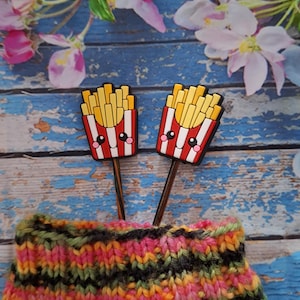 Könnte beinhalten: Zwei skurrile Stricknadelaufsätze in Form von Pommes Frites mit lächelnden Gesichtern, vor einem blauen Holzgrund. Die Aufsätze sind rot und weiß mit gelben Pommes. Ein farbenfrohes, gestreiftes Strickstück befindet sich im Vordergrund.