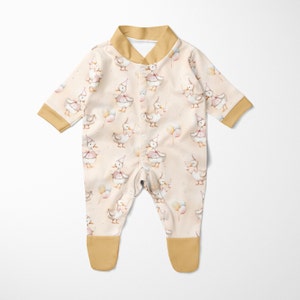 May include: A cream-coloured baby onesie with a mustard yellow collar and cuffs. The onesie features a repeating pattern of watercolour ducks wearing party hats and holding balloons.