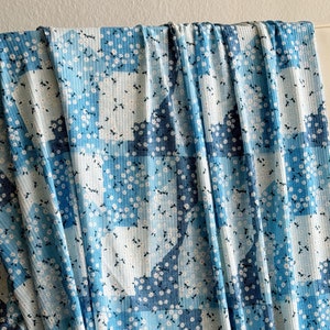 May include: Blue fabric with a floral pattern. The fabric features a patchwork design with varying shades of blue and white, adorned with small white flowers and green leaves. The fabric is draped, showing its texture and flow.