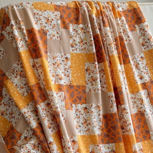 May include: A patchwork fabric with a repeating pattern of squares in shades of orange, yellow, and beige. Some squares feature floral prints, while others have paisley or striped designs. The fabric is draped over a white surface.