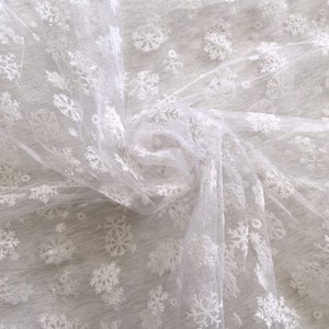 May include: White sheer fabric with snowflake embroidery. The fabric is draped and folded, showing the intricate details of the snowflake design.