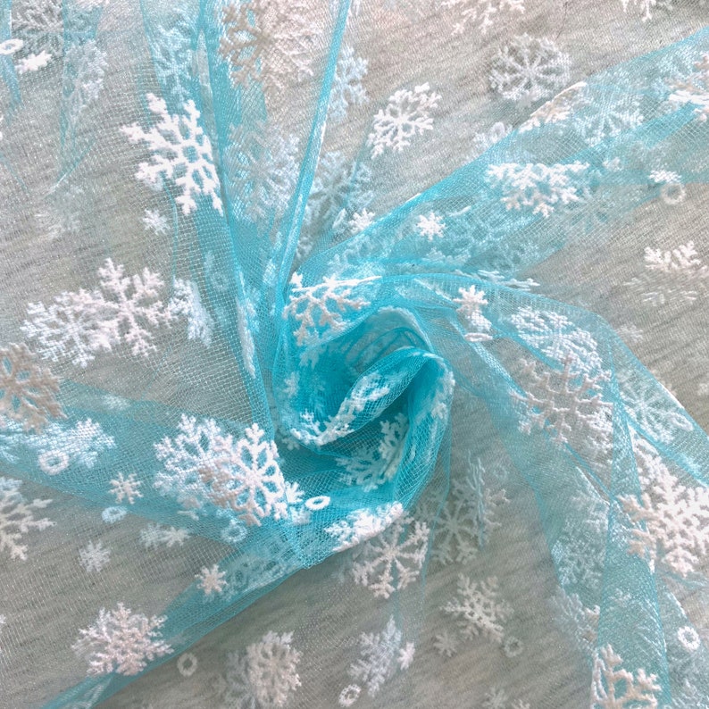 May include: Light blue tulle fabric with white snowflake embroidery. The fabric is gathered and twisted, showing the texture of the tulle.
