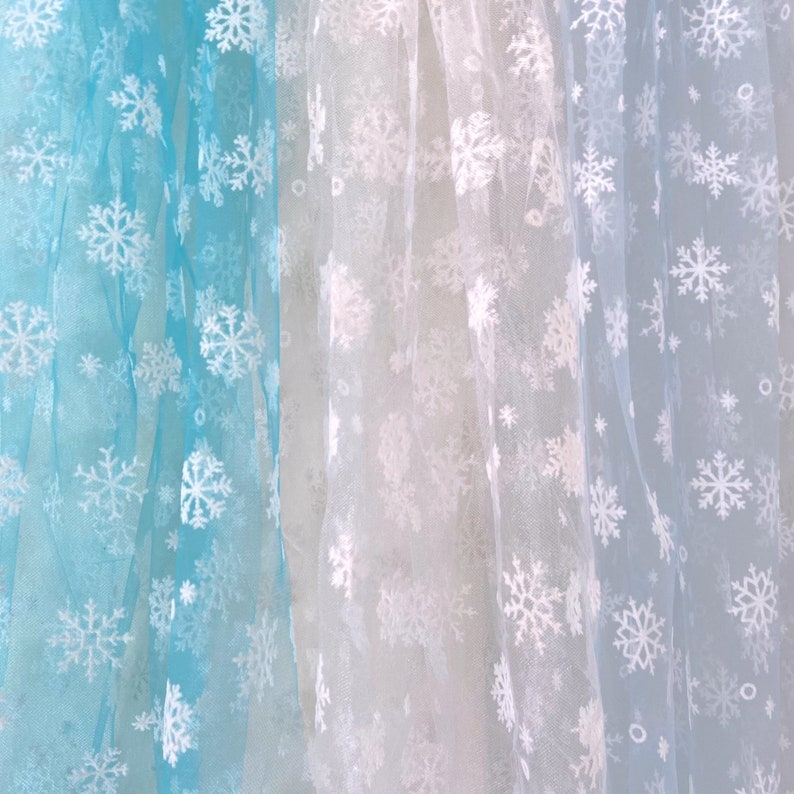 May include: Three sheer fabric panels with snowflake patterns. The panels are light blue, white, and a lighter shade of blue.