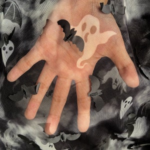 May include: A hand with a ghost and bats printed on a black mesh fabric. The fabric has a repeating pattern of ghosts and bats.
