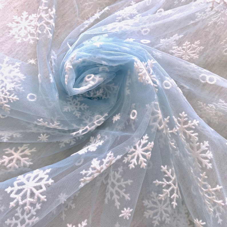 May include: Light blue tulle fabric with white snowflake embroidery. The fabric is gathered and draped, showing the delicate texture of the tulle and the intricate detail of the snowflakes.