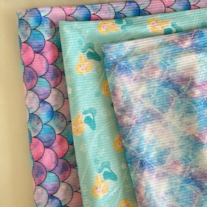 May include: Three fabric swatches with a mermaid theme. The top swatch is a light blue with a white and pink floral pattern. The middle swatch is a light blue with a white and pink floral pattern and a mermaid silhouette. The bottom swatch is a pink and blue watercolor pattern with a mermaid scale design.