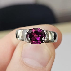 May include: A silver ring featuring a large, oval-cut, deep magenta gemstone. The gemstone is set in a simple, modern band. The ring is held up against a blurred background, showcasing the jewelry's design and color.