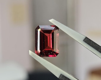 May include: A deep red, emerald-cut gemstone held by a pair of metal tweezers. The stone features a rectangular shape with faceted edges that reflect light, displaying a rich, saturated crimson hue throughout its transparent, polished surface.