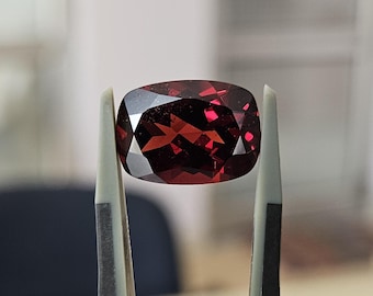 May include: A deep red, cushion-cut gemstone with a faceted surface that reflects light. The stone is held by a pair of grey tweezers with black rubberized tips, showcasing the clarity and rich crimson color of the polished, transparent mineral.