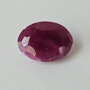 May include: Oval-shaped ruby gemstone with a deep red hue. The faceted surface reflects light, creating a sparkling effect. The gemstone is set against a white background, highlighting its color and clarity.