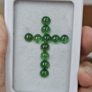 May include: A collection of ten small, round, translucent green gemstones arranged in the shape of a cross. The stones are displayed inside a rectangular white plastic case with a soft, light gray felt lining that holds them securely in place.