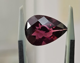 May include: A pear-shaped, deep magenta-colored gemstone with a faceted surface. The stone is held between two light gray tweezers, showcasing its translucent quality and geometric cut that reflects light across its polished, angular surfaces.