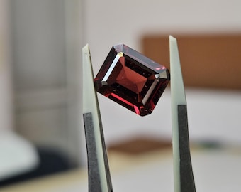May include: A deep red, emerald-cut gemstone held by a pair of grey-tipped metal tweezers. The stone features a faceted surface with sharp, clean edges and a transparent, polished finish that reflects light across its rectangular, symmetrical shape.