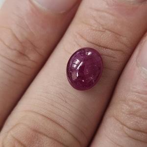 Könnte beinhalten: Ein tief magentafarbener, ovaler Edelstein mit einem glatten, polierten Cabochon-Schliff. Der Stein weist eine durchscheinende Qualität mit sichtbaren inneren Einschlüssen und einer reflektierenden Oberfläche auf, die einen satten rot-violetten Farbton zeigt.