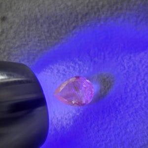 May include: A pear-shaped, translucent pink gemstone illuminated by a handheld ultraviolet light source. The gemstone exhibits a distinct glow under the UV light, highlighting its faceted surface and clear, light pink coloration in a focused beam.