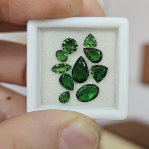 May include: A collection of ten loose, faceted green gemstones displayed in a square white plastic case with a white foam insert. The stones vary in shape, including pear, oval, round, marquise, and triangular cuts, all featuring a deep green hue.