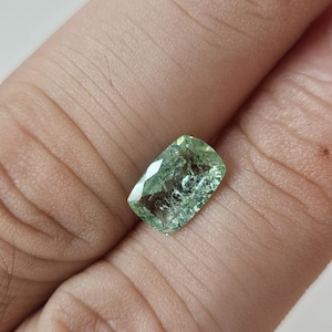 May include: A cushion-cut gemstone with a light green hue and visible internal facets. The stone features a rectangular shape with rounded corners and a polished surface that reflects light, showcasing a translucent appearance and natural inclusions.