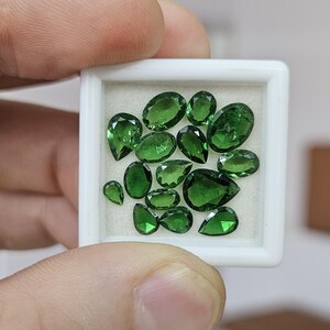 May include: A collection of small, faceted green gemstones in various shapes, including pear, oval, and round cuts, arranged inside a square white display box with a light-colored foam insert to hold the loose stones securely in place.