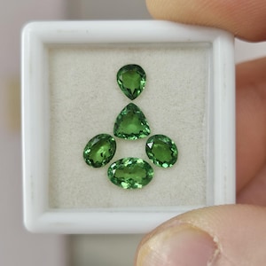 May include: A collection of five faceted green gemstones arranged in a white square display box. The set includes one pear-shaped stone, one triangular stone, and three oval-shaped stones, all featuring a vibrant, translucent emerald-green hue.