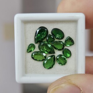 May include: A collection of ten loose, faceted green gemstones displayed in a small, square white plastic box with a cream-colored velvet insert. The stones vary in shape, including pear, oval, and round cuts, all exhibiting a deep, vibrant green hue.