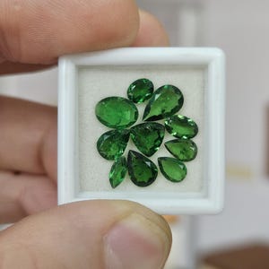 May include: A collection of ten loose, faceted green gemstones in various shapes, including pear, oval, and triangular cuts, arranged inside a small, square white display box with a white foam insert for secure storage and presentation.
