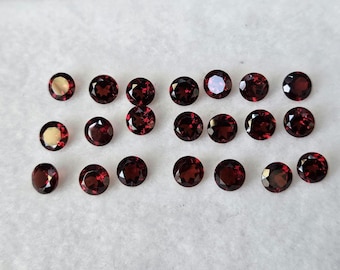 May include: A collection of round, deep red gemstones arranged on a white surface. The gemstones vary slightly in size and have a polished, reflective surface, showcasing their rich color and clarity. These gems are likely garnets.