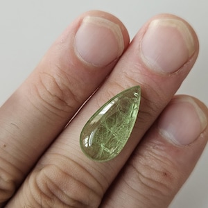 May include: A pear-shaped, translucent light green gemstone with visible internal needle-like inclusions. The stone features a smooth, polished surface with a faceted top, displaying a bright, pale lime color throughout its teardrop-shaped body.