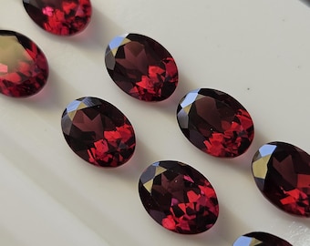 May include: A collection of oval-cut, deep red gemstones. The gems are arranged on a white surface, showcasing their faceted surfaces and internal brilliance. The color ranges from dark crimson to lighter shades.