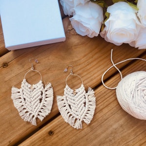 Puede incluir: Un par de pendientes de macramé blanco con detalles de flecos y cuentas doradas. Los pendientes están colgados sobre una superficie de madera.