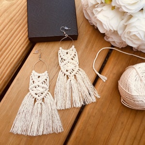 May include: A pair of white macrame earrings with fringe. The earrings are hanging from a wooden surface.