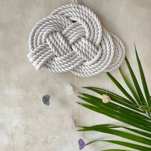 May include: A white rope wall hanging in the shape of a cloud with four crystals hanging from it. The crystals are clear, green, pink, and purple.