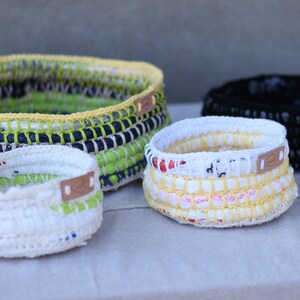 May include: Five crocheted baskets made from recycled materials in various sizes and colors, including white, green, yellow, and black. Each basket has a leather tag.