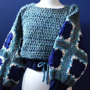 May include: A handmade, blue crochet sweater with square patterned sleeves. The sweater features a drawstring waist and a round neckline. The sleeves have a patchwork design with shades of blue and white.