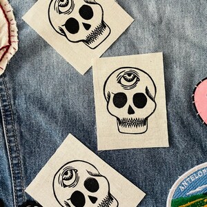 Puede incluir: Tres parches cuadrados de tela con diseños de calaveras negras. Cada calavera tiene cuencas oculares grandes, una mandíbula detallada y un símbolo de ojo sobre la frente. Los parches están sobre un fondo de mezclilla, para ropa.