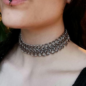 Könnte beinhalten: Ein silbernes Choker-Halsband aus Kettengeflecht. Die Halskette besteht aus kleinen, ineinandergreifenden Metallringen, die einen geschichteten Effekt um den Hals erzeugen. Der Choker ist ein auffälliges Accessoire, das einen gotischen oder alternativen Stil widerspiegelt.
