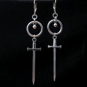 Può includere: Un paio di orecchini color argento con un design a spada. Ogni orecchino presenta una piccola sfera, un cerchio e un ciondolo a spada. Gli orecchini sono sospesi su uno sfondo nero.